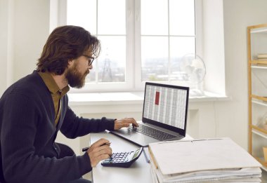 Ofis masasında oturan, dizüstü bilgisayarda çalışan ve hesap makinesi kullanan bir adam. Ofisteki modern bir dizüstü bilgisayarın elektronik iş dosyalarıyla meşgul bir muhasebeci.