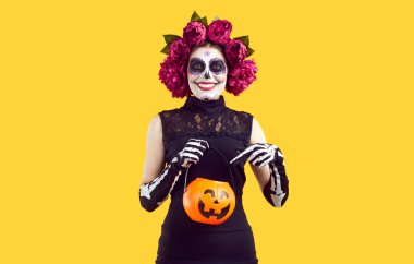 Cadılar Bayramı 'nda şeker toplamak için balkabağı sepeti tutan yaratıcı kılık değiştirmiş kadın. Meksika tarzı giyinmiş kafasında çelenk olan kız turuncu arka planda bir kova dolusu işaret ediyor.