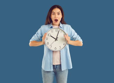 Shocked stunned wondered brunette woman in casual clothes blue shirt, jeans and white t-shirt is holding clock in her hands on blue background. Wasting time, late, empty life, crisis concept. clipart