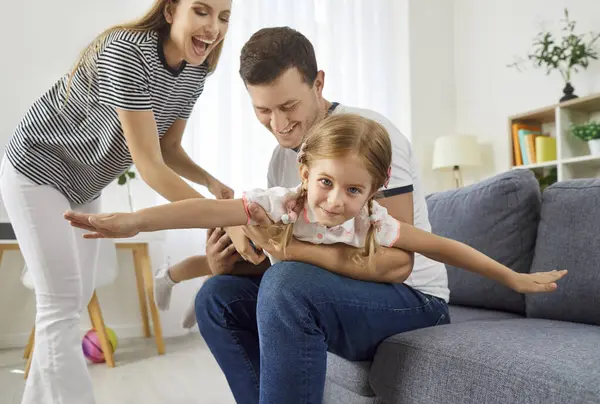 Mutlu genç ebeveynler kızlarıyla vakit geçiriyorlar, birlikte eğleniyorlar, uçan uçak oynuyorlar. Neşeli baba, kollarını uçak gibi kaldıran gülümseyen küçük kızı kaldırıyor. Aile eğlence zamanı..