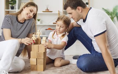 Neşeli aile birlikte vakit geçiriyor, ebeveynler ve küçük kızları tahta küplerle eğitici oyunlar oynuyor, takım olarak çalışıyor, oyuncak insan figürleriyle kule inşa etmek için blokları diziyorlar..