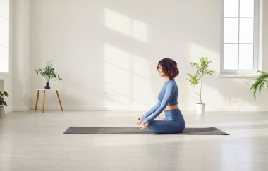 Yogi sportif çekici kadın yoga yapıyor, Padmasana egzersizi yapıyor, Mudra jesti ile Lotus pozu veriyor spor kıyafetlerini giyip beyaz yoga stüdyosunda, evde, yan bakışta mindere oturuyor.