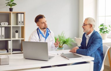 Ciddi bir erkek doktor ya da hemşire, yaşlı bir erkek hastaya danışıyor, hastane ofisinde bir masada oturuyor. Tıbbi konsültasyon sırasında konuşuyorlar ve bakım sağlıyorlar..