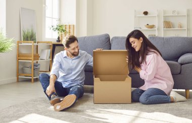 Mutlu, genç, neşeli bir çift evde paketleri açıyor, oturma odasında yerde oturuyor ve gülümsüyor açık karton kutuya bakıyor. Teslimat, online alışveriş, paket konseptini al.