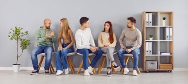 Adaylar iş görüşmesini beklerken konuşuyor. Şirket çalışanları iş toplantısının başlamasını bekliyor. Bir grup insan ofis koridorunda raf ve saksı bitkisiyle otururken sohbet ediyorlar.