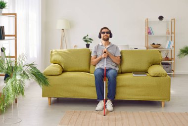 Çubuk değnekli genç bir adamın portresi evde kanepede oturmuş kulaklıklarla müzik dinliyor ya da sesli kitap dinliyor. Koyu gözlüklü ve kanepeye uzanmış bir erkek..