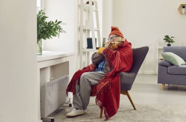 Donmuş kıdemli adam evdeki kış soğuğuyla savaşıyor, dış giysilere bürünmüş ve battaniyeye sarılmış. Isıtıcının varlığına rağmen, oda soğuk bir rahatsızlık hissi taşıyor..