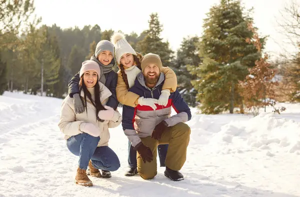 Çocukların kış parkında bir arada durup kameraya baktığı mutlu bir aile. Karlı ormanda dışarıda yürüyen iki çocuklu genç ebeveynler. Aile eğlencesi ve kış tatili konsepti.