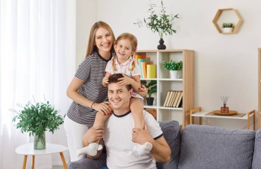 Evdeki kameraya bakan mutlu aile portresi. Mutlu bir baba oturma odasında koltukta oturuyor, küçük kızı omuzlarında ve gülümseyen annesi yanlarında duruyor ve kızına sarılıyor..