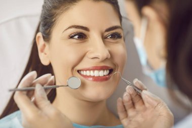 Modern diş hekimliği kliniğinde gülümseyen güzel bir kadının dişlerini diş aletleriyle muayene edip tedavi eden genç bir dişçi. Diş sağlığı, hijyen ve tıp konsepti.