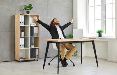 Zaferin ve işte başarının ifadesi. Mutlu iş adamı başarısına sevinir ve iyi haber onuruna yumruklarını kaldırır. Genç adam işyerinde oturuyor ve zaferini kutluyor..