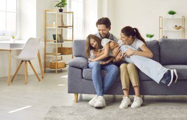 Aile eğlencesi. Sevgi dolu ebeveynler, evde eğlenirken küçük kızlarına sataşır ve gıdıklarlar. Komik Kafkasyalı genç baba, anne ve kızları oturma odasında kanepede birlikte vakit geçiriyorlar.. 