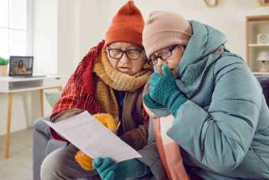 Kış kıyafetleriyle evdeki oturma odasında oturan yaşlı bir çiftin portresi ve battaniyeye sarılmış elektrik faturasına bakmak. Isıtma sorunları konsepti.