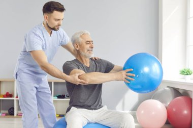 Koç ya da sağlık çalışanı olgun bir adamın spor egzersizleri yapmasına yardım ediyor. Spor salonunda formda top oynuyor. Fizyoterapist rehabilitasyon merkezindeki yaşlı erkek hastalara yardım ediyor..