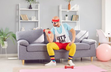 Spor giyimli komik bir genç adam, oturma odasındaki kanepede oturan halterlerle evde egzersiz yapıyor. Erkek kişi evde egzersiz yapıyor. Spor, spor, sağlık ve evde eğitim kavramı.