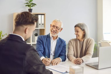 Yaşlı çift emeklilik planları ve yatırım stratejilerini finans danışmanıyla tartışıyor. Zarif giyimli, neşeli gülüşlü yaşlı bir çift ofiste masa sözleşmesi imzalıyor..