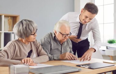 Genç bir erkek danışman ya da erkek bir finans danışmanıyla birlikte ofiste oturan kendine güvenen gri saçlı yaşlı çift sağlık sigortası ya da yatırımlarla ilgili bir sözleşme imzalıyor..