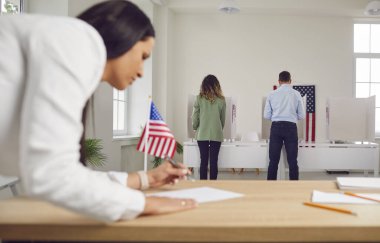 Oy verme merkezinde çalışan genç bir kadın. Amerikan seçmenlerinin oy verme kabininde arka arkaya oy kullanması ve oy verme kabininde ABD bayraklarının olması. Seçim günü ve demokrasi kavramı.
