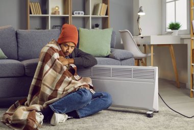 Elektrikli ısıtıcının yanında battaniyeyle kaplanmış hayal kırıklığına uğramış bir adam oturuyor. Sıcak giyinmiş bir adam, kendini ısıtma kaloriferinde ısıtırken evde üşüyor. Isıtma sorunları, güç krizi kavramı