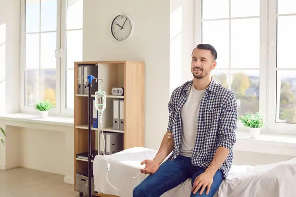 stock image Portrait of a young happy smiling man sitting on the couch in medical clinic and receiving IV drip infusion and vitamin therapy in his blood. Cheerful smiling person receiving injection therapy.