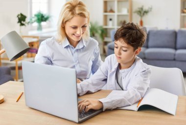 Mutlu anne oğluna ödevlerinde yardım ediyor evde online e-öğrenim için dizüstü bilgisayar kullanıyor. Aile bağları, iç ortamdaki öğrenim oturumunda eğitim ve destek yoluyla güçlenir.