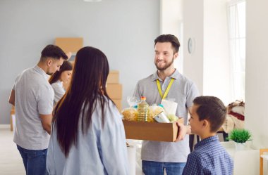 Happy gönüllü, sosyal yardım merkezindeki mülteci bir aileye bir kutu yiyecek veriyor. Bu sahnede kamu hizmeti, insani yardım ve ihtiyacı olanlara yardım çabaları vurgulanıyor.