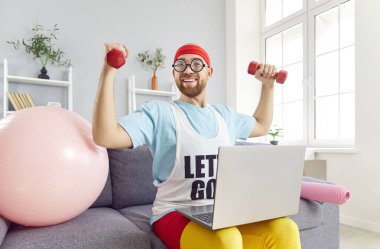 Spor kıyafetli komik bir genç, kameraya bakıyor. Antrenörü takip eden dambıllar kullanıyor. Evdeki kanepede oturuyor. Uzaktan antrenman, spor ve mizah konsepti.