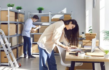 Çevrimiçi mağazada çalışan insanlar. Kadın ve erkek işçiler, küçük bir depoda, raflarda ve masalarda karton kutularla envanter kontrolü yapıyorlar. E ticaret KOBİ işletmesi