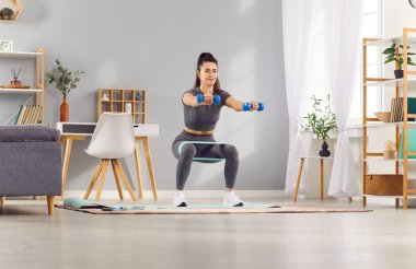 Kadın spor salonuna gidiyor, spor yapmaya odaklanıyor, çömelme hareketleri yapıyor ve direnç bandıyla egzersiz yapıyor. Etkili bir egzersiz yaptığı için spor ve antrenman çok açık..