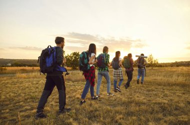 Bir grup genç turist sırt çantalarıyla sahada yürüyorlar. Mutlu insanlar, erkek ve kadınlar gün batımında doğa yürüyüşüne çıkıyorlar. Macera, seyahat ve turizm konsepti.
