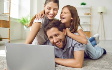 Mutlu, neşeli anne, baba ve çocuk evde oturma odasında dinleniyor, dizüstü bilgisayar kullanıyor, TV programlarını izliyor, komik şakalara gülüyor, birlikte eğleniyorlar.