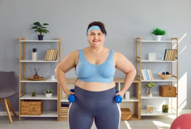 Gülümseyen şişman kadın evde elinde dambıllarla spor egzersizleri yapıyor. Artı kilolu kız kameraya neşeli görünüyor. Spor, sağlıklı yaşam tarzı ve vücut pozitifliği.