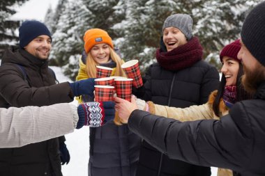 Happy smiling friends relaxing, clinking a glass of hot coffee among snow covered trees in snowy winter park outdoors. Young people in winterwear having fun in forest drinking hot beverage. clipart