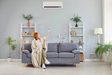 Battaniyeye sarılı bir kız, klimayı açıp kapatıyor, sıcak hava odası sıcaklığını kontrol ediyor, uygun performansı ayarlıyor, ev ortamını ayarlıyor, soğuk havada ideal iklim ısıtma sistemi sağlıyor.