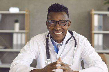 Mutlu Afro-Amerikan doktor, kameraya dostça bir ifadeyle bakıyor. Laptop bilgisayar ekranı kamera görüntüsü. Beyaz laboratuvar önlüğü ve gözlüklü gülümseyen siyah adamın portresi.