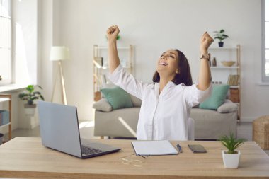 Zor projeyi bitiren kadın kendini mutlu hissediyor. Neşeli iş kadını iyi yapılmış bir işin memnuniyetini yaşar. Kadın dizüstü bilgisayarla masada oturuyor, ellerini kaldırıyor ve gülümsüyor.
