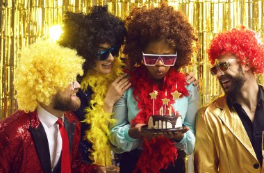 Parti kutlaması ve komik aktiviteler. Disko saç perukları ve şenlikli kostümlü mutlu arkadaşlar doğum günü kadınını tebrik edin. Parlak gece kulübünün arka planında pasta mumları üfleyin.