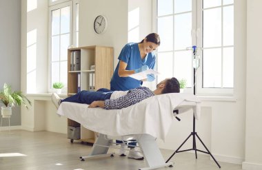 Genç bayan hemşire ya da doktor randevu ile rapor tutuyor ve klinikte kanepede yatan erkek hastasına danışmanlık yapıyor ve serum ve vitamin tedavisi görüyor..