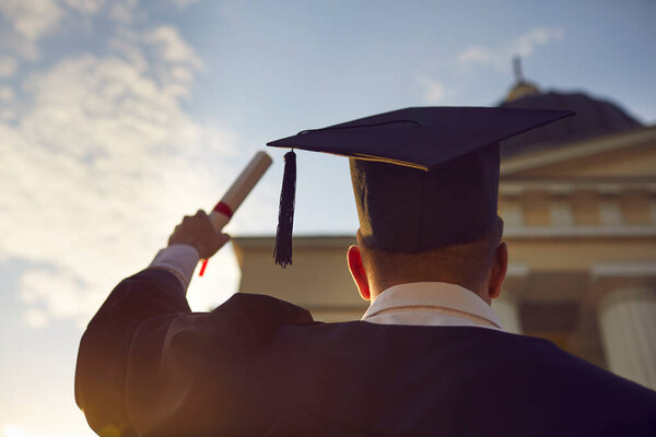 Выпускная церемония. Студент мужского пола в традиционной одежде и шляпе с кисточкой, стоящей с поднятым дипломом на фоне заката и здания университета на размытом фоне. Нижний вид сзади