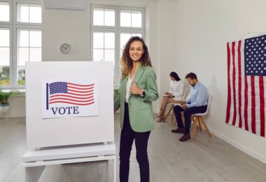 Mutlu gülümseyen Amerikalı seçmen kadının portresi kameraya neşe içinde bakıyor. Seçim günü oy verme kabininde kağıt oy pusulasıyla işaret veriyor. ABD bayrakları seçim yapıyor..