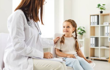 Pediyatrist, muayene sırasında ya da klinikte konuşurken mutlu bir kız hastayla konuşur. Doktor, çocuk için hastane ziyareti sırasında dostane bir atmosfer sağlayarak tıbbi bakım sunuyor..