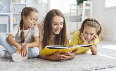 Genç mutlu anne, iki küçük tatlı çocuğuyla birlikte oturma odasında yerde uzanmış kitap okuyor. Gülümseyen kadın çocuklarla vakit geçiriyor. Aile eğlencesi kavramı..
