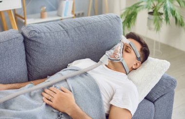 Yüzünde oksijen maskesi olan genç bir adam kanepede uyuyor, yakın plan. Astım tedavisi, evde rehabilitasyon. Kronik oksijen eksikliği, terapi uykusu, aile ilaçları, apne.
