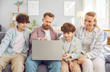 Çocuklu mutlu bir aile, evdeki bir kanepede dinleniyorlar, online bir eğlenceyle meşguller. Bir dizüstü bilgisayar kullanmak, modern eğlenceyi sembolize etmek ve internetin aile bağları ve rahatlama için kullanılması.