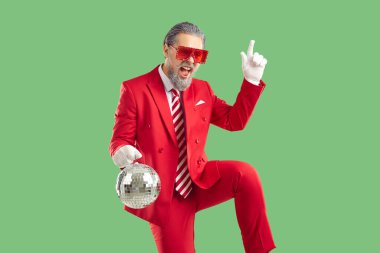 Grey-haired old man wearing red suit and white gloves, Santa style. He dancing with disco ball in hands on green background. He pointing his index finger up. Celebrating Christmas, New Year concept. clipart