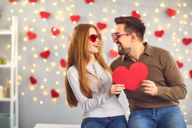 Sevgi dolu bir çift ellerinde aşkın kırmızı bir sembolünü tutuyorlar ve birbirlerine bakıyorlar. Erkek ve kız, arka planda asılı duran çelenk ve kalpler üzerinde kırmızı şenlikli gözlüklerle eğleniyorlar..