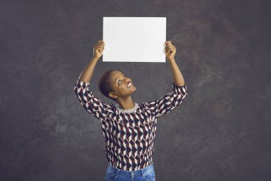 Mutlu gülümseyen genç siyah kadın gri arka planda yalnız duran boş beyaz kağıt pankart gösteriyor. Stüdyo portresi. Güzel, kısa saçlı, Afrikalı bir kadın. Elinde temiz, boş bir maket levhası var.