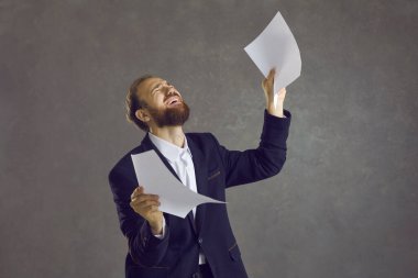 Gri arka planda genç beyaz bir adam mutlu ve başarılı hissederek kağıtları kaldırdı. Zafer konsepti. Mavi ceketli başarılı adam.