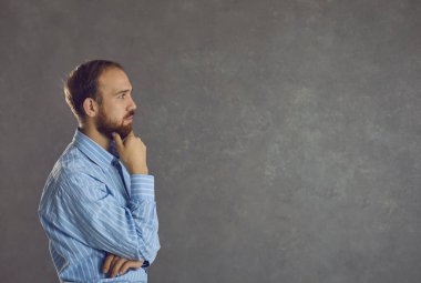 Çenesini tutan dalgın bir ifadeyle kafası karışmış ciddi bir iş adamının profil görüşü. Gömlekli yetişkin sakallı bir adam gri arkaplandaki yazı için boş alana bakıyor. Yansıma kavramı.
