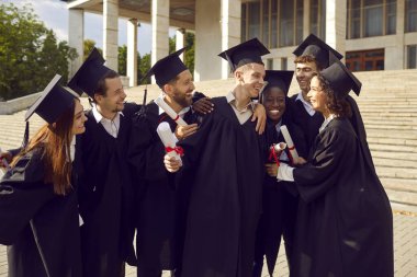 Bir grup gülümseyen, mutlu, çok ırklı uluslararası mezun öğrencinin portresi. Üniversite mezuniyet elbisesi içinde kucaklaşıp eğleniyorlar ve diplomalarını açık havada tutuyorlar. Eğitim ve mezuniyet kavramı.